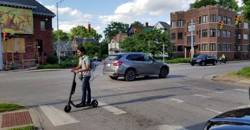 Man crossing a crosswalk riding a scooter.